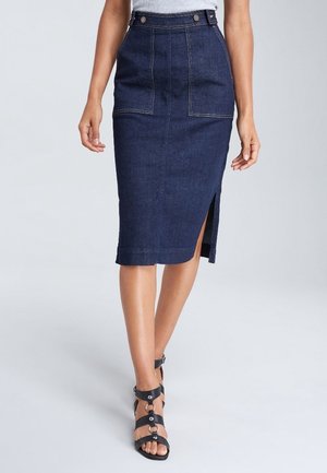 Femme portant une jupe midi en denim bleu foncé avec poches avant et fente latérale, associée à des sandales noires à talons hauts à lanières.