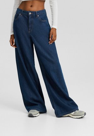 Jean denim bleu foncé à jambes larges avec une taille haute, doté de poches avant et de surpiqûres contrastées, associé à des baskets gris clair.