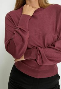 Woman wearing a long-sleeve, ribbed cuff mauve sweater with one arm crossed and the other hand near her chin.
