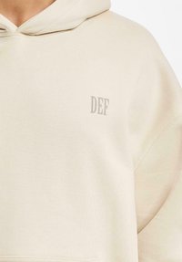 Lys beige hettegenser i myk stoff med kengurulomme, som har en liten grå "DEF"-logo på brystet og en avslappet passform.