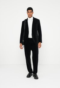 Čierny kordurový blazer s jednou vreckom, kombinovaný s čiernymi nohavicami a bielou košeľou na gombíky. Čierne topánky dotvárajú outfit.