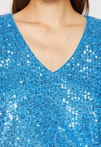 Top blu in paillettes con design a V. Caratterizzato da una texture scintillante con paillettes ravvicinate, che creano una superficie riflettente.