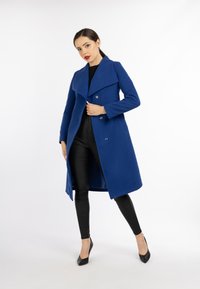 Palton din lână albastră, cu revere late, design cu două rânduri de nasturi și buzunare laterale. Asortat cu pantaloni negri, ajustați, și tocuri înalte negre.