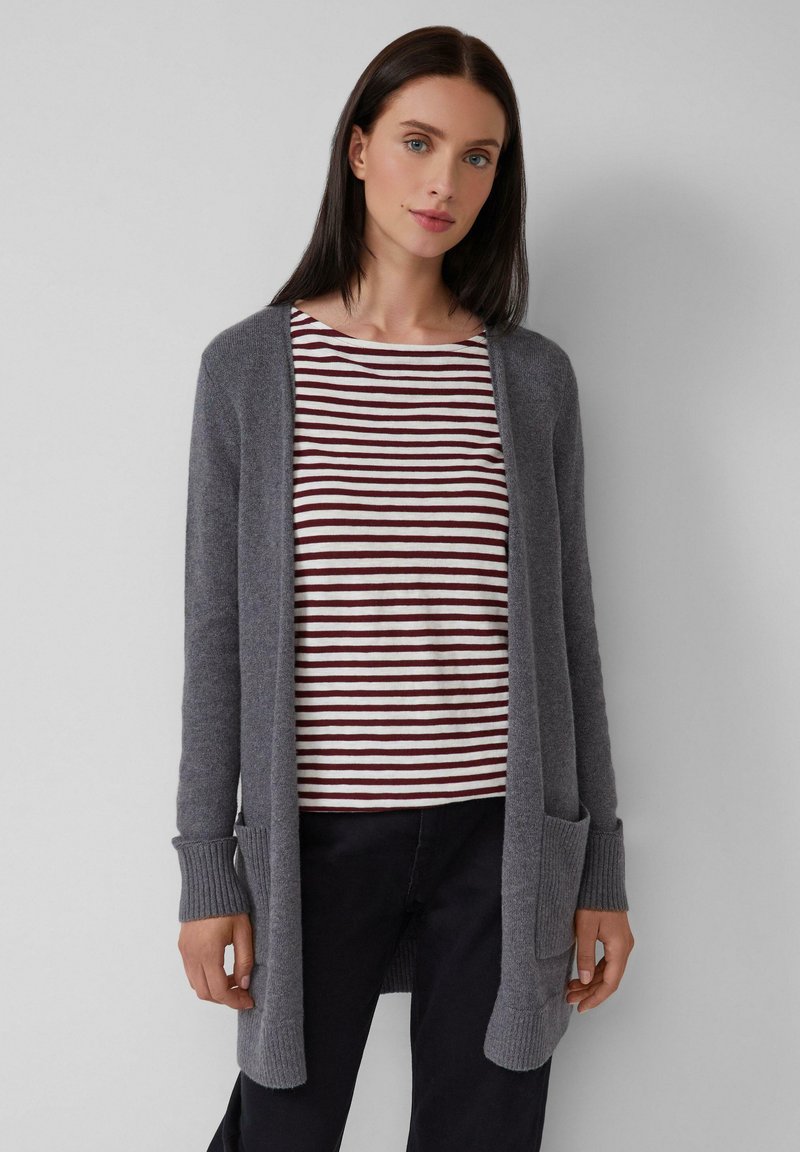 Mujer vistiendo un cardigan gris abierto sobre una camiseta blanca y burdeos a rayas y pantalones negros, de pie contra un fondo claro y liso.