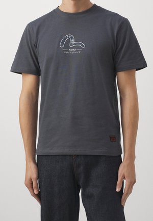 Hombre con camiseta de manga corta gris oscuro con un logo estilizado blanco y texto en japonés en el pecho, y vaqueros azul oscuro.