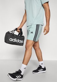 Μαύρη τσάντα τύπου duffel με λευκό λογότυπο «adidas» και χερούλια μεταφοράς. Το μοντέλο φοράει ανοιχτοπράσινο αθλητικό πουκάμισο, ταιριαστό σορτς, μαύρα παπούτσια και λευκές κάλτσες.