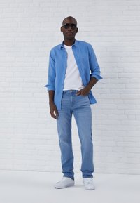 Light blue button-up shirt over a white t-shirt, paired with light blue jeans and white sneakers. Dark sunglasses complete the look.