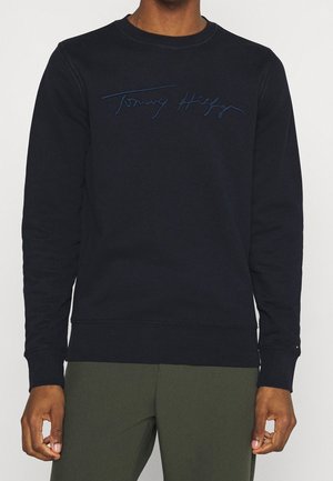 Mężczyzna noszący czarną długą bluzę z haftowanym napisem „Tommy Hilfiger” na klatce piersiowej, zestawioną z oliwkowozielonymi spodniami.