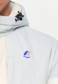Light grey jacket featuring a textured white sherpa collar, a K-WAY logo with blue and red accents, and a black toggle closure.