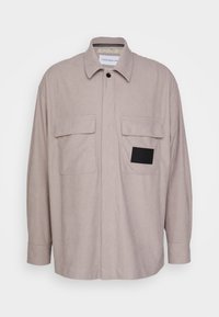 Giacca camicia grigio chiaro con motivo a quadri, chiusura con bottoni, due tasche sul petto e dettaglio in patch nero sulla tasca sinistra.