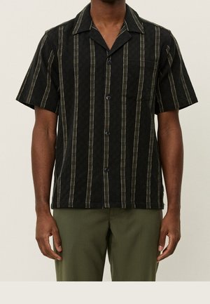 Homme portant une chemise noire à manches courtes avec des rayures beiges verticales et un pantalon vert olive sur un fond uni.