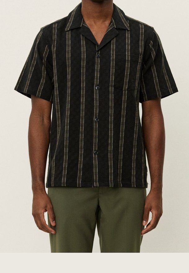 Homme portant une chemise noire à manches courtes avec des rayures beiges verticales et un pantalon vert olive sur un fond uni.