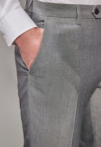 Pantaloni gri, croiți, dintr-o țesătură netedă, cu buzunare laterale și o croială slim, asociați cu manseta unei cămăși albe cu nasturi.