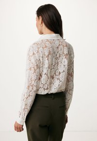 Camicia bianca in pizzo con motivo floreale e maniche lunghe, dotata di un colletto bianco. Abbinata a pantaloni verde oliva a vita alta.