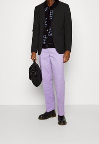 Černé sako, vzorovaný černý kardigan, světle lila kalhoty a černé kotníkové boty. Drží černou kabelku, stojí na neutrálním pozadí.