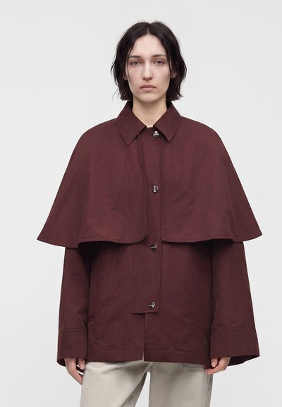Personne aux cheveux courts et foncés portant une veste cape marron foncé ample à boutons sur un pantalon beige clair devant un fond blanc uni.