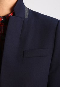 Blazer bleu marine à finition texturée, avec un col gris contrastant et une poche poitrine unique. Chemise à carreaux discrète en dessous.