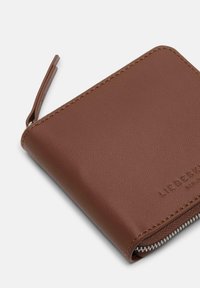 Brązowy skórzany portfel o gładkiej fakturze, z zamkiem błyskawicznym i jasnobrązowym przeszyciem. Z przodu znajduje się tłoczone logo.