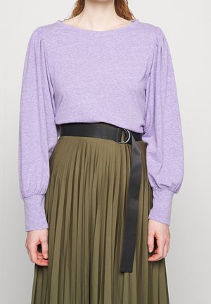 Frau trägt eine langärmelige, lavendelfarbene Bluse, die in einen olivgrünen, plissierten Rock mit einem schwarzen Gürtel mit einer baumelnden Lasche gesteckt ist.