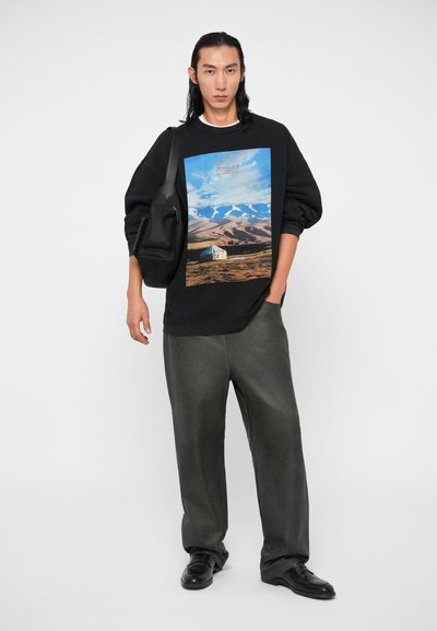 Felpa nera con grafica paesaggistica, abbinata a pantaloni grigi larghi e scarpe nere. L'outfit presenta una vestibilità morbida e un design minimalista.