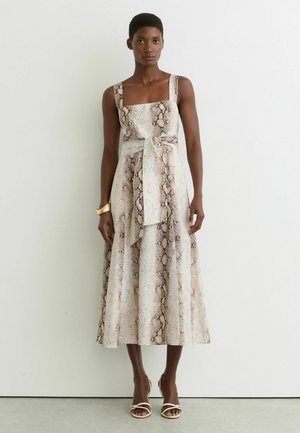 Frau trägt ein ärmelloses, midi-langes Kleid mit Schlangenprint, breiten Trägern und Frontknoten, kombiniert mit weißen Sandalen mit Absatz und einem goldenen Armband.