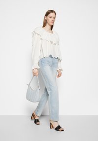 Valkoinen röyhelöblusa, jossa on rei'itettyjä yksityiskohtia, vaaleansiniset suorat farkut, mustat korkokengät ja vaaleansininen päällystetty käsilaukku.