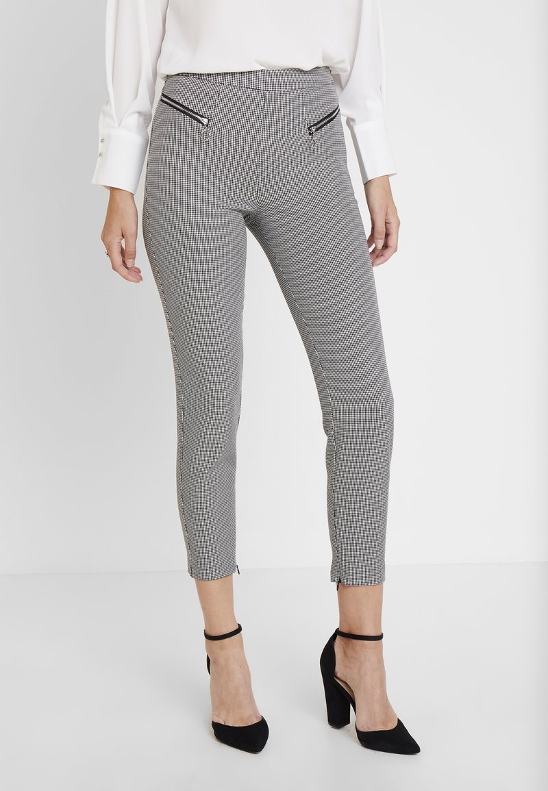 Femme portant un pantalon slim noir et blanc à carreaux avec des poches zippées à l'avant et des chaussures noires à talons hauts avec bride à la cheville.
