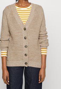 Beige stickad cardigan med V-ringning, fem knappar och ribbade ärmar över en gult och vitt randig topp. Strukturerad tyg och avslappnad passform.