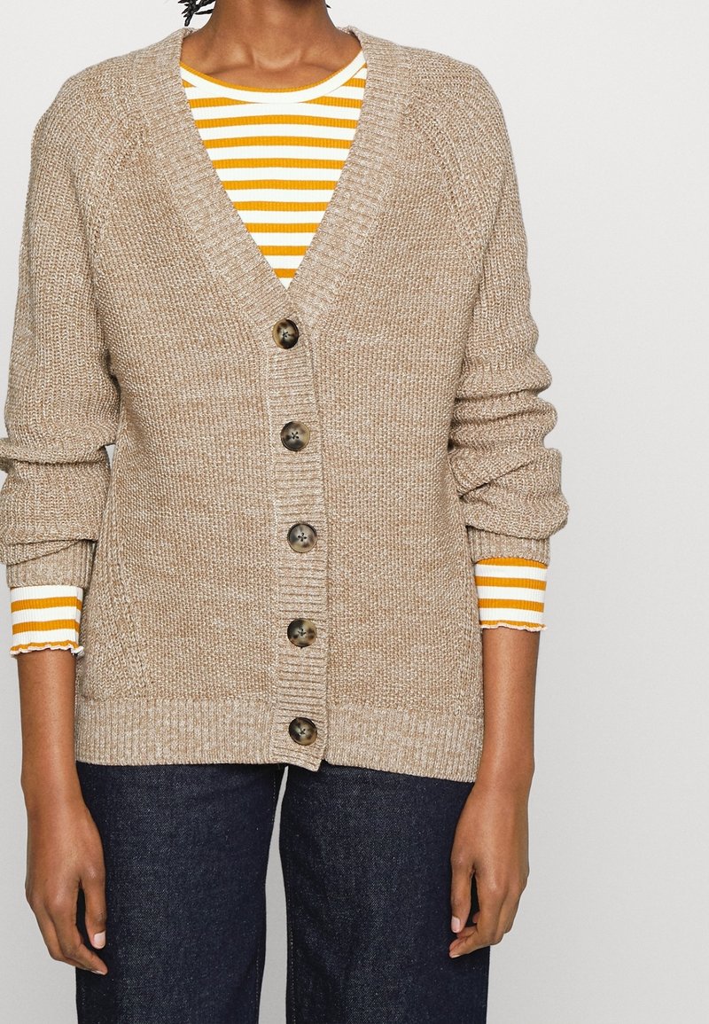 Beige stickad cardigan med V-ringning, fem knappar och ribbade ärmar över en gult och vitt randig topp. Strukturerad tyg och avslappnad passform.