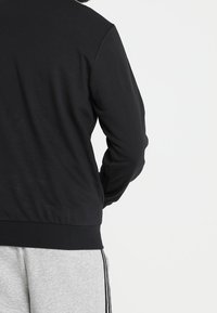 Veste noire à manches longues en tissu lisse, avec un ourlet et des poignets côtelés, vue de l'arrière, portée avec un pantalon de sport gris.