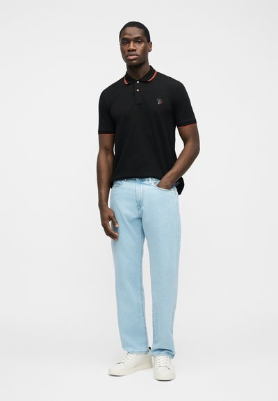 Άνδρας που φοράει μαύρο πόλο μπλουζάκι με κόκκινη διακόσμηση, ανοιχτό μπλε τζιν και λευκά αθλητικά παπούτσια, στέκεται με το ένα χέρι στην τσέπη μπροστά σε λευκό φόντο.