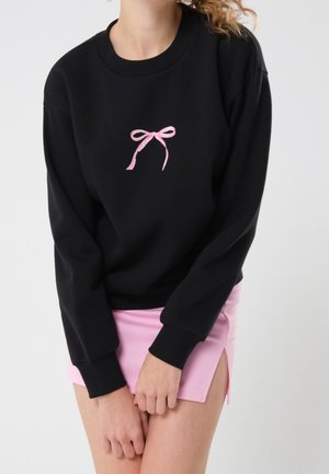 Femme portant un sweat-shirt noir avec un motif de nœud rose et une mini-jupe rose clair avec une fente latérale, debout les mains jointes.