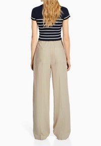 Kvinna med långt blont hår som bär en svart och vit randig kortärmad topp och högmidjade beiga vida byxor, sedd bakifrån.