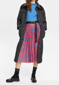 Svart vadderad kappa över en blå ribbad topp, i kombination med en plisserad kjol med röda och blå virvlande mönster, svart bälte och skinande svarta skor.