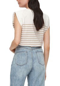 Žena s dlouhými tmavými vlasy, oblečená v bílém topu bez rukávů se béžovými pruhy a světle modrých džínech s vysokým pasem, viděná ze zad.