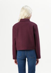 Veste courte bordeaux avec un col, en tissu texturé, et manches longues, associée à un jean bleu, vue de dos.