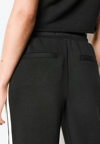Pantaloni neri con una superficie liscia, dotati di due tasche posteriori e una vestibilità comoda. La cucitura laterale presenta una striscia bianca a contrasto.