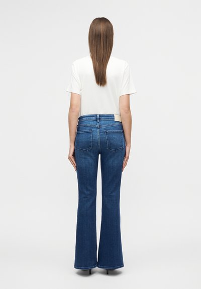 Παντελόνι τζιν καμπάνα βαθύ μπλε με ψηλή μέση, πέντε τσέπες και ίσια ραφή. Συνδυασμένο με απλό λευκό T-shirt.