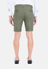 Pantaloni corti khaki con vestibilità slim e orli arrotolati, dotati di due tasche posteriori con chiusura a bottone, indossati con scarpe da ginnastica nere.