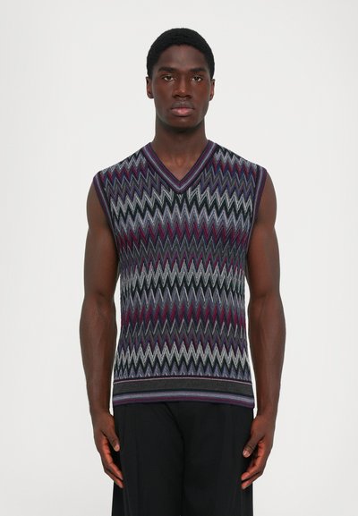 Mężczyzna w bezrękawnikowym swetrze V-neck w purpurowym, czarnym, szarym i białym wzorze zygzaka, stojący na tle jasnego, jednolitego tła.