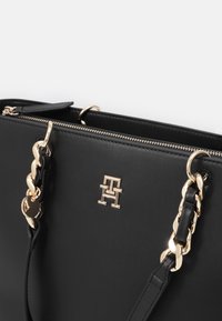 Black leather handbag with gold hardware, featuring a chain accent on the straps and a logo prominently displayed on the front.
