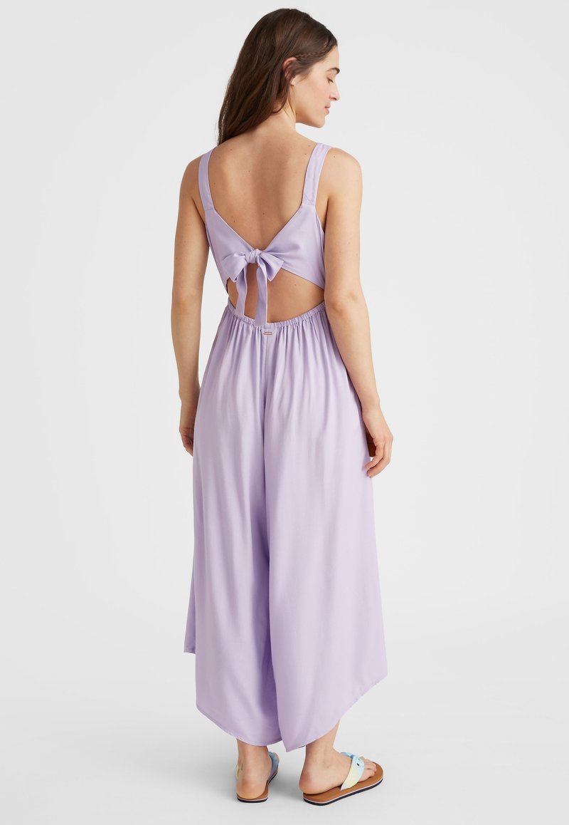 Lila Jumpsuit mit weitem Bein, offener Rückseite mit einer Schleife und elastischem Taillenbund. Das Modell trägt Flip-Flops mit pastellfarbenen Akzenten.