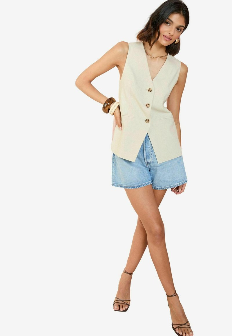Chaleco sin mangas color crema con escote en V y tres botones, combinado con pantalones cortos de denim claros y sandalias negras con tiras.