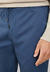 Pantalones azul marino de tejido suave, con un cordón ajustable con puntas de metal y bolsillos laterales. Suéter beige texturizado visible encima.