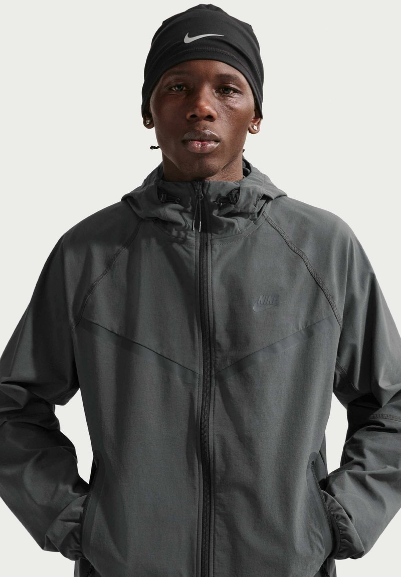 Man wearing black Nike beanie and dark gray Nike zip-up jacket with hands in pockets, facing forward against plain light background.