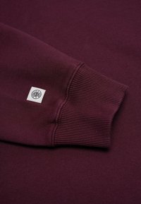 Burgundy-coloured collegepaita, jossa on ribbaus hihansuissa. Sen etuosassa on pieni logo-merkki kontrastina olevaan valkoiseen neliöön. Kangas on sileää ja pehmeää.
