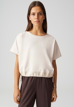 Donna che indossa una maglia bianca sporca, a maniche corte e ampia, con coulisse regolabili all'orlo e pantaloni marrone scuro, in piedi davanti a uno sfondo grigio.