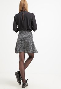Blusa di raso nera con apertura a chiave sulla schiena, abbinata a una gonna a campana in maglia grigia. La modella indossa collant trasparenti e scarpe nere lucide.