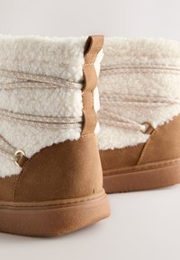 Bottes en suédine camel avec une encolure en shearling blanche, présentant des coutures décoratives et une semelle en caoutchouc texturée. Lacets élastiques pour un ajustement réglable.