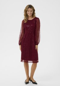 Rochie burgundă din dantelă, cu mâneci lungi transparente, brodată cu paiete, cu un tiv până la genunchi și un model texturat. Pur tată cu tocuri negre ascuțite.